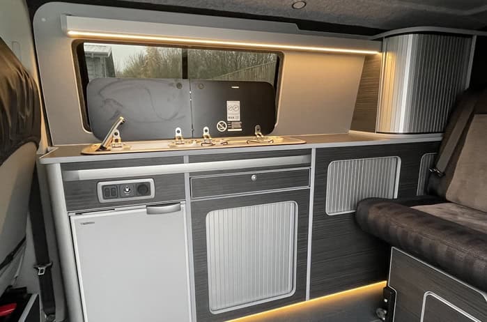 Ford Transit Custom Campervan Kitchen Unit dark grey cabinetry with integrated sink, dual gas hob, fridge, and warm LED strip lighting inside van interior.