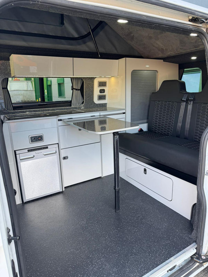 Ford Transit Custom Campervan Kitchen Unit Bespoke in white gloss with black seating and table, fully fitted inside a van with rubber flooring and open sliding door background.