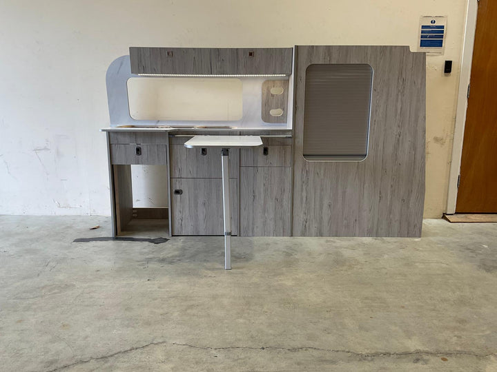 Ford Transit Custom Campervan Kitchen Unit Bespoke in gloss white and charcoal two-tone design with fold-out table and side tambour door, displayed on a concrete floor with plain wall background.