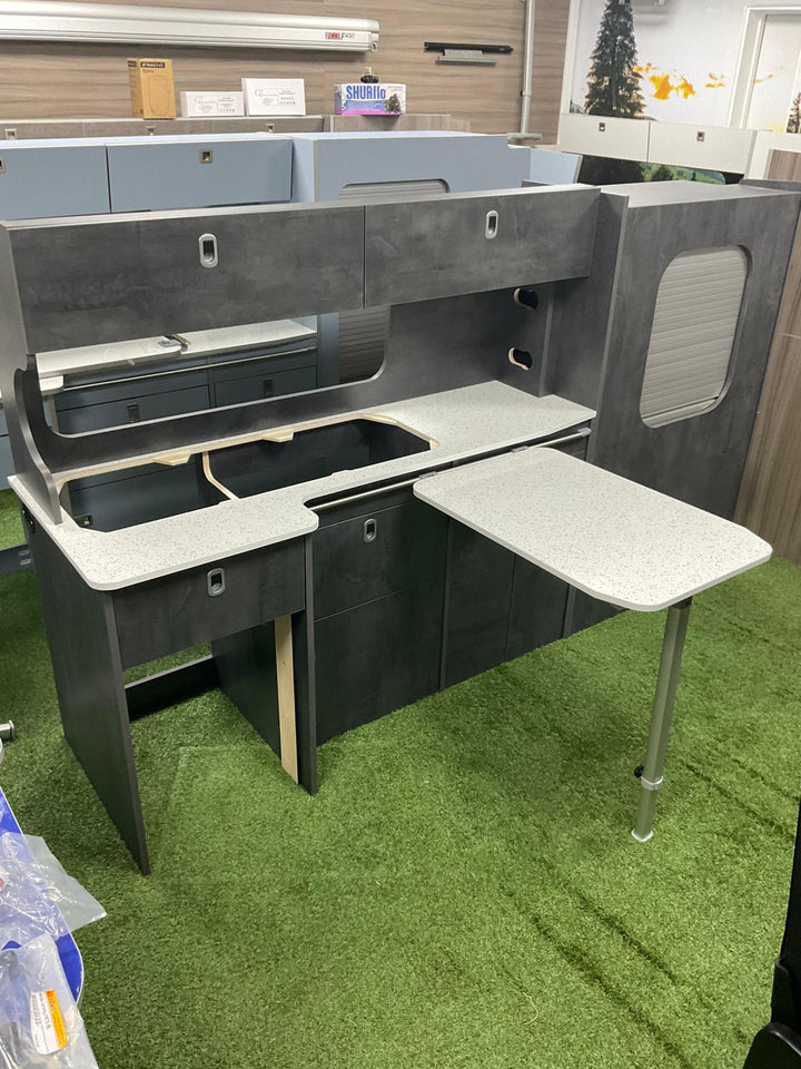 Ford Transit Custom Campervan Kitchen Unit Bespoke in light grey wood-effect finish with overhead lockers and fold-out table, displayed in a workshop on a concrete floor against a plain wall background.