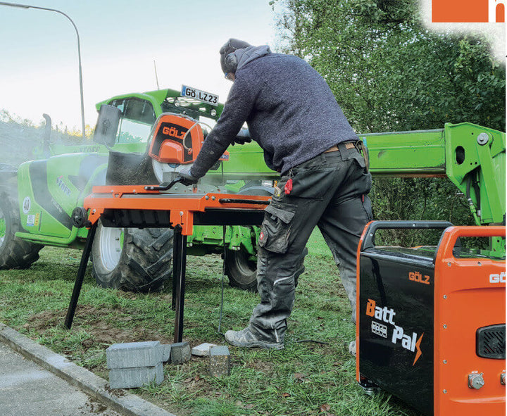 A  worker operates a masonry saw powered by the GÖLZ BattPak 5048 on a grassy worksite, with a green telehandler vehicle in the background.