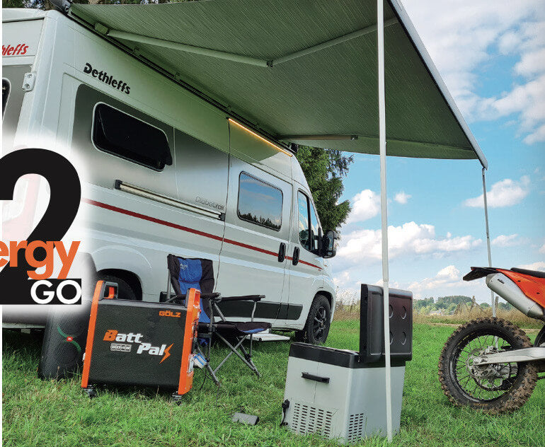 The GÖLZ BattPak 5048 supplies electricity to camping appliances under an awning beside a white camper van, with a motorbike and chairs arranged for outdoor leisure.