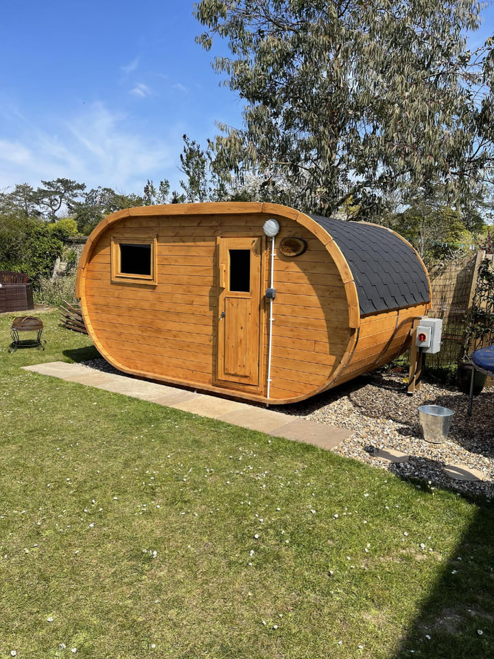 InMedias Oval Sauna with honey-colored wood and black roof shingles on a backyard lawn with clear blue sky 