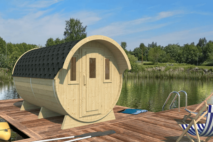 A wooden barrel-shaped camping cabin with a black shingled roof placed on a wooden dock by a calm lake, surrounded by green trees under a bright blue sky.