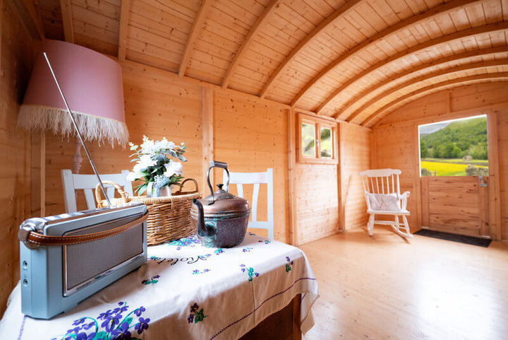 Interior of an InMedias Traditional Shepherd Hut with wooden walls and arched ceiling, a table with a floral cloth, a kettle, chairs, and an open door showing a green landscape, on a bright natural background.