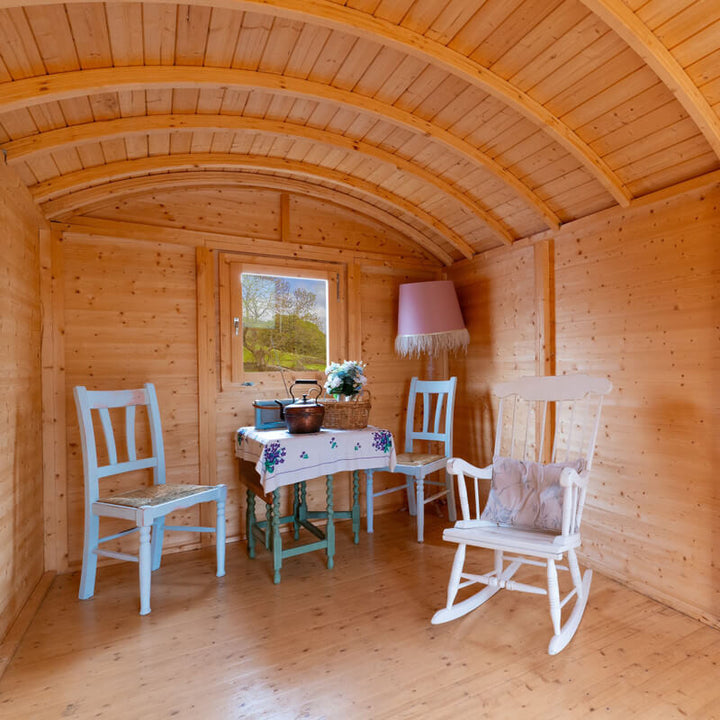 Cosy wooden interior of an InMedias Traditional Shepherd Hut with curved ceiling, pastel chairs, table, and a pink lamp, on a bright natural background.