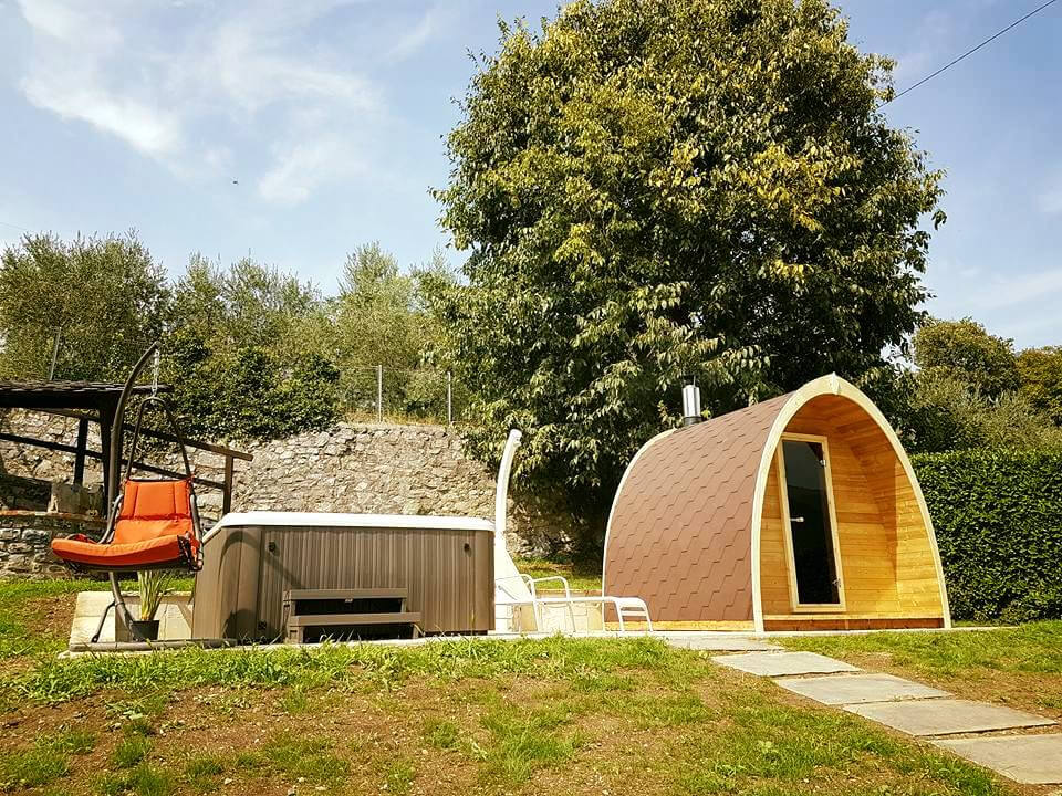 InMedias Sauna Pod 300 beside a hot tub and lounge chairs in a backyard garden on a sunny day with blue sky and trees.