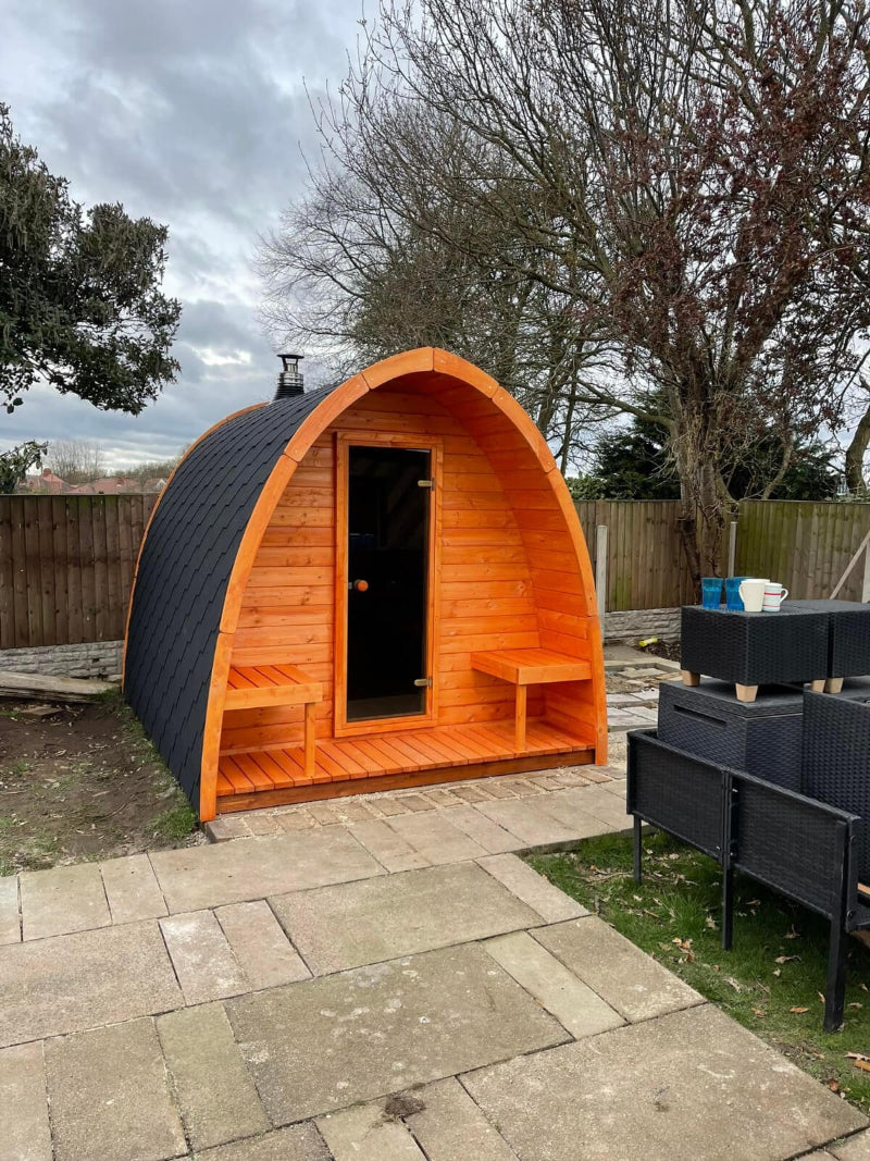 InMedias Sauna Pod 300 in vibrant orange wood finish standing on paving slabs on a cloudy gray sky background.

