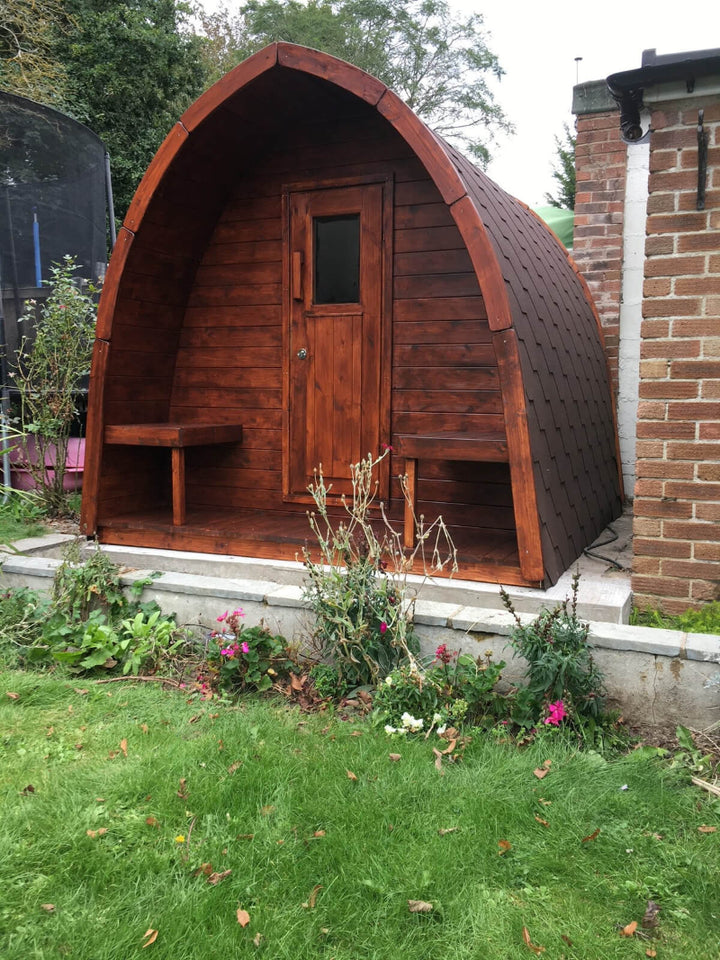 InMedias Sauna Pod 300 with rich brown wood and square window door placed in a garden on a natural green grass background.