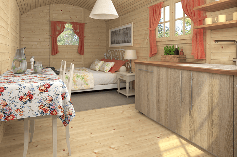 Interior view of a luxury InMedias Traditional Shepherd Hut fitted with a kitchen, dining area, and a bed with red curtains and wood furnishings, on a bright natural background.