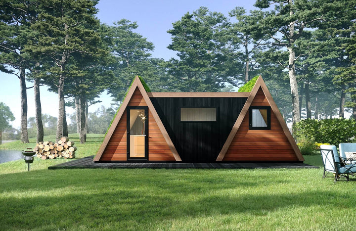 Front view of double A-frame glamping cabin with green roofs and mixed timber cladding, surrounded by trees, lake, and outdoor seating.