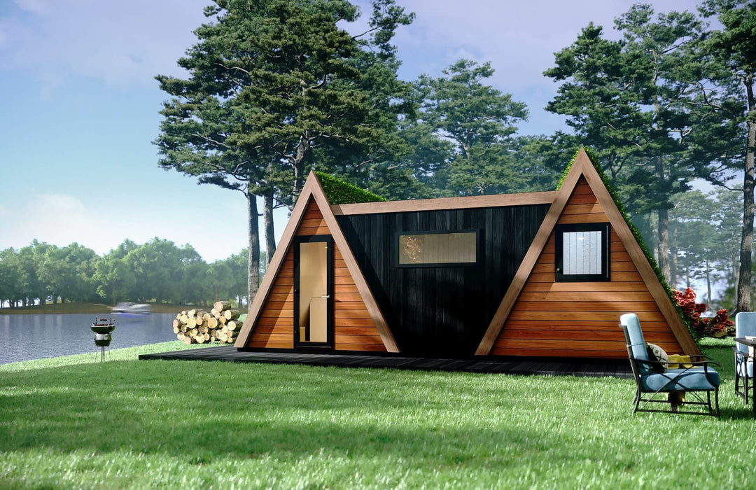 Twin A-frame glamping pod with black cladded middle section, lakeside view and BBQ grill setup in a lush outdoor setting.
