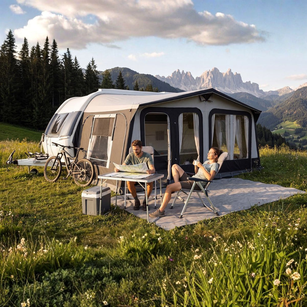 A large pop-up caravanserail tent is pitched in a grassy mountain meadow with two people sitting outside at a small table, bicycles parked nearby, and mountains and pine trees in the background.