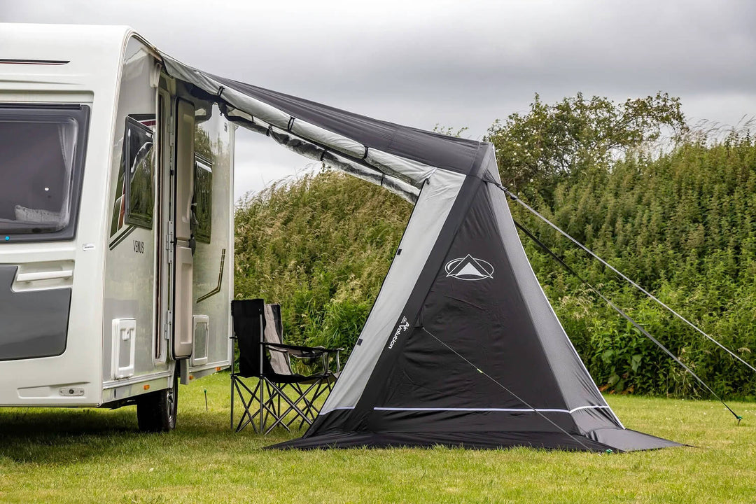 Side view of a black and grey SunnCamp Air 325 awning with a sloping roof and support poles, attached to a white caravan beside green hedges on a grassy campsite.