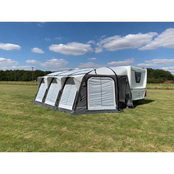 A Sunncamp Icon Air Awning attached to a white caravan, displaying four connected sections with black support beams and white panels, set on green grass under a blue sky with white clouds