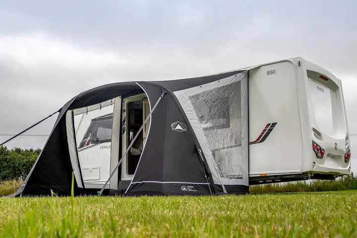 SunnCamp Swift Air 325 caravan awning with black and grey fabric, clear window panels and an open entrance, attached to a white Venus caravan on a grassy campsite under a cloudy sky.