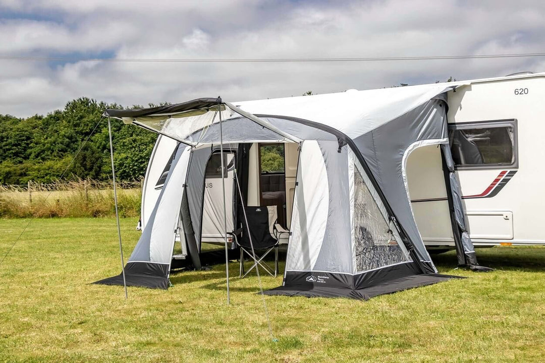 SunnCamp Swift Air 220 SC awning with an open front canopy, transparent side windows, and sturdy support poles attached to a white caravan, set on a green lawn beside hedges.