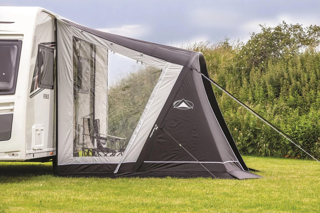 Side view of a SunnCamp Swift Air 325 awning with a large transparent window panel, dark fabric walls and guy ropes, attached to a white caravan beside tall green hedges.