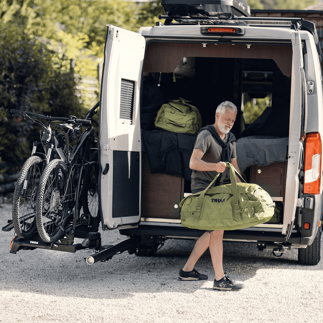 Man unloading gear beside van equipped with Thule Veloswing towbar on an outdoor campsite background.