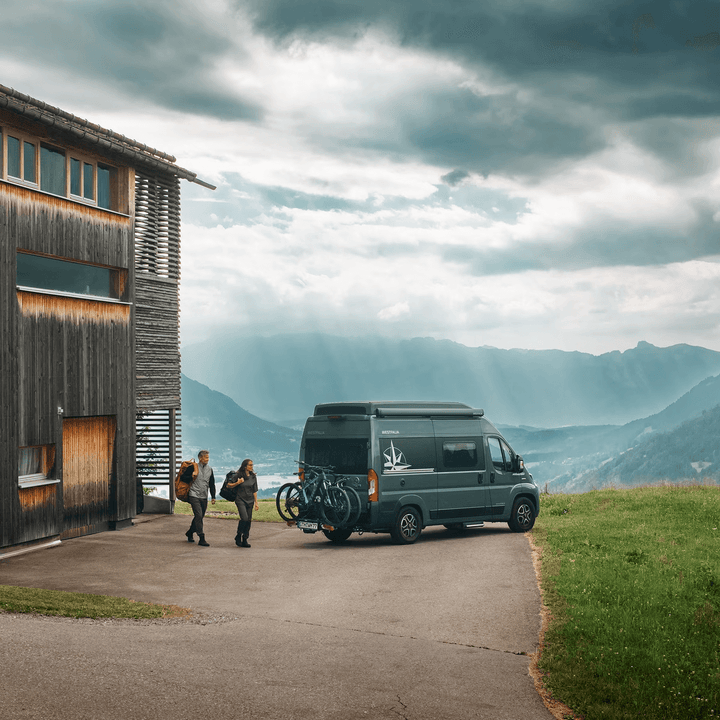 Van with bikes mounted on Thule Veloswing towbar parked near mountains on an outdoor background.
