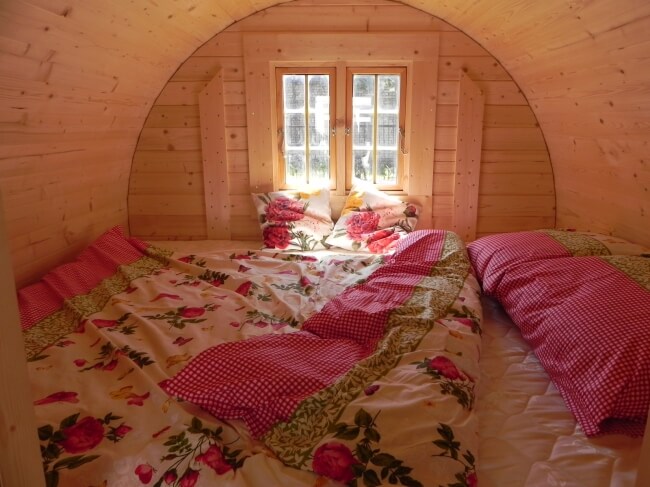 Cozy bedroom inside a Viking Industrier 2.2 x 4m Sleeping Barrel with wooden interior, floral blankets, and a window with sunlight streaming in.
